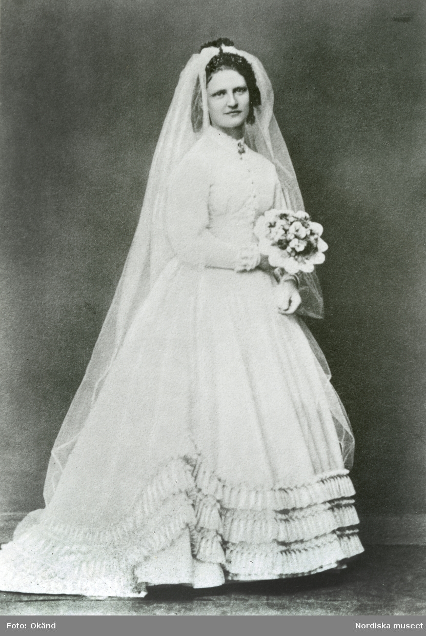 Bröllopsfoto Kristinehamn. Porträtt i helfigur av bruden prostinnan Sophie Lindström (född Lagergren) iklädd brudklänning med snäv ärm och volanger nedtill.
