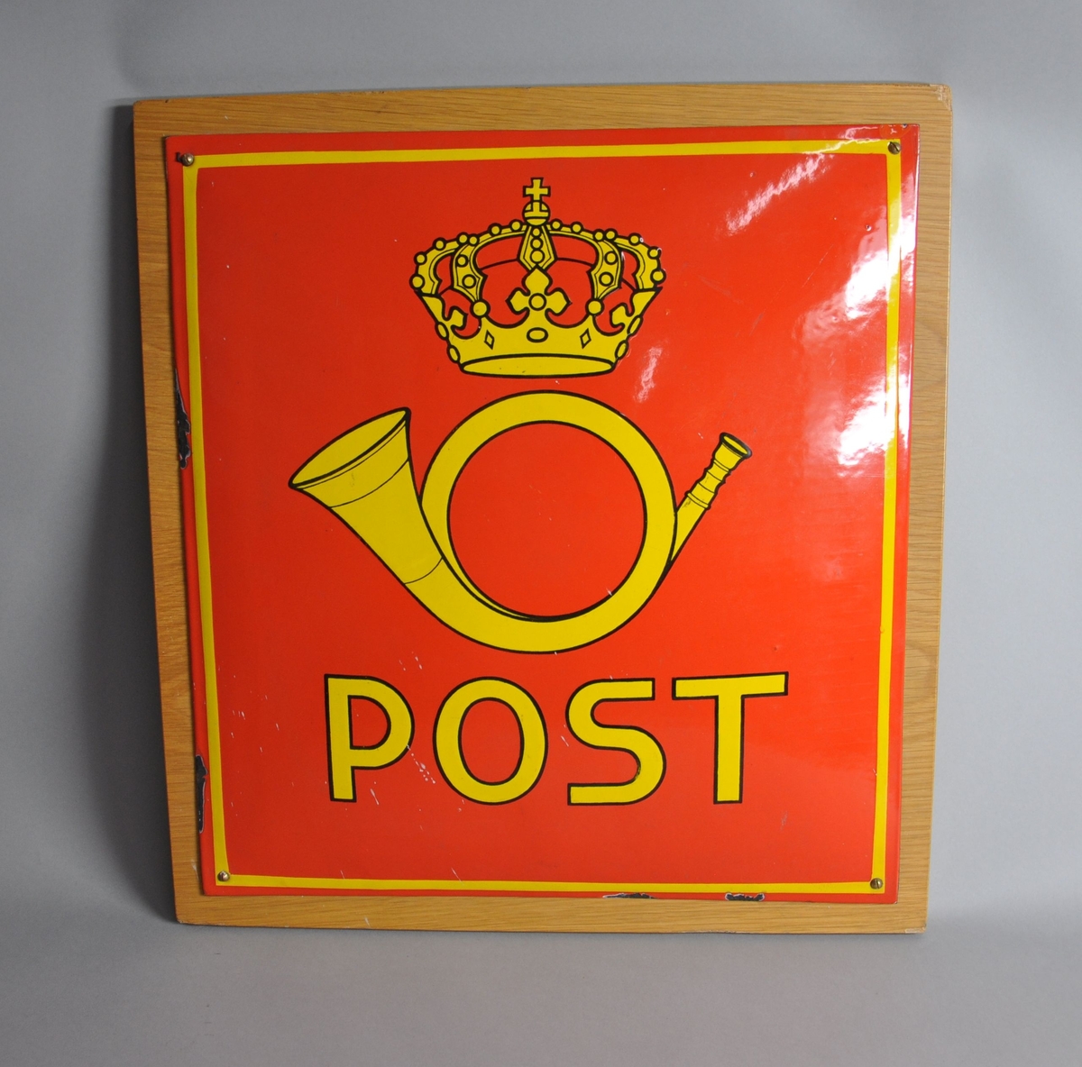 Rødt postskilt av metall på treplate. Gul innskrift og dekor, emblem fra det norske postvesen.