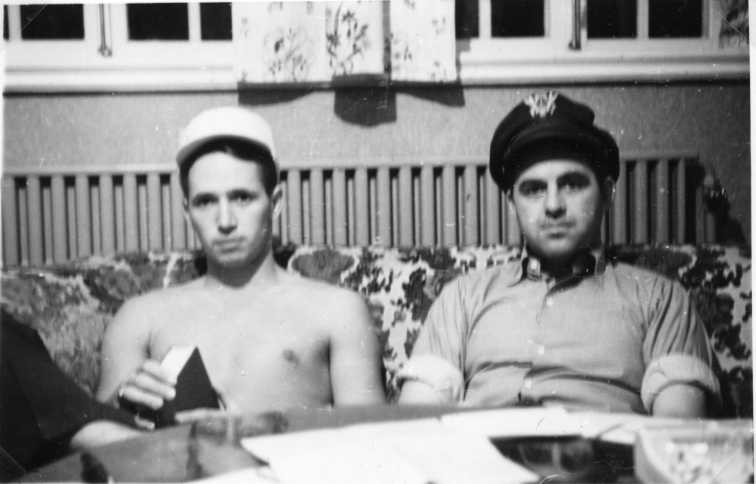 Två män sitter i blommig soffa, En med bar överkropp och keps den andra med uppkavlade skjortärmar och uniformsmössa.
