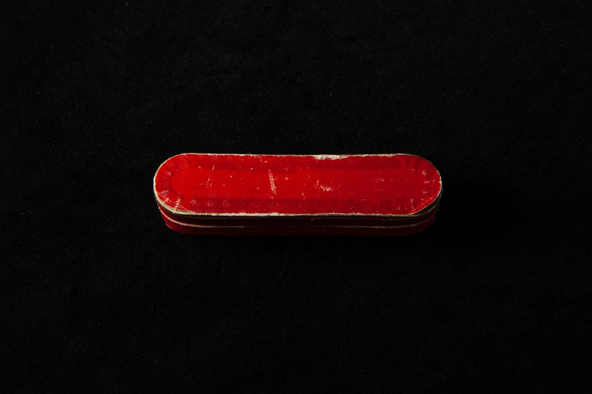 Ovalt formad pappask med lock. Röd med guldkant. På insidan finns en etickett med påskrift.