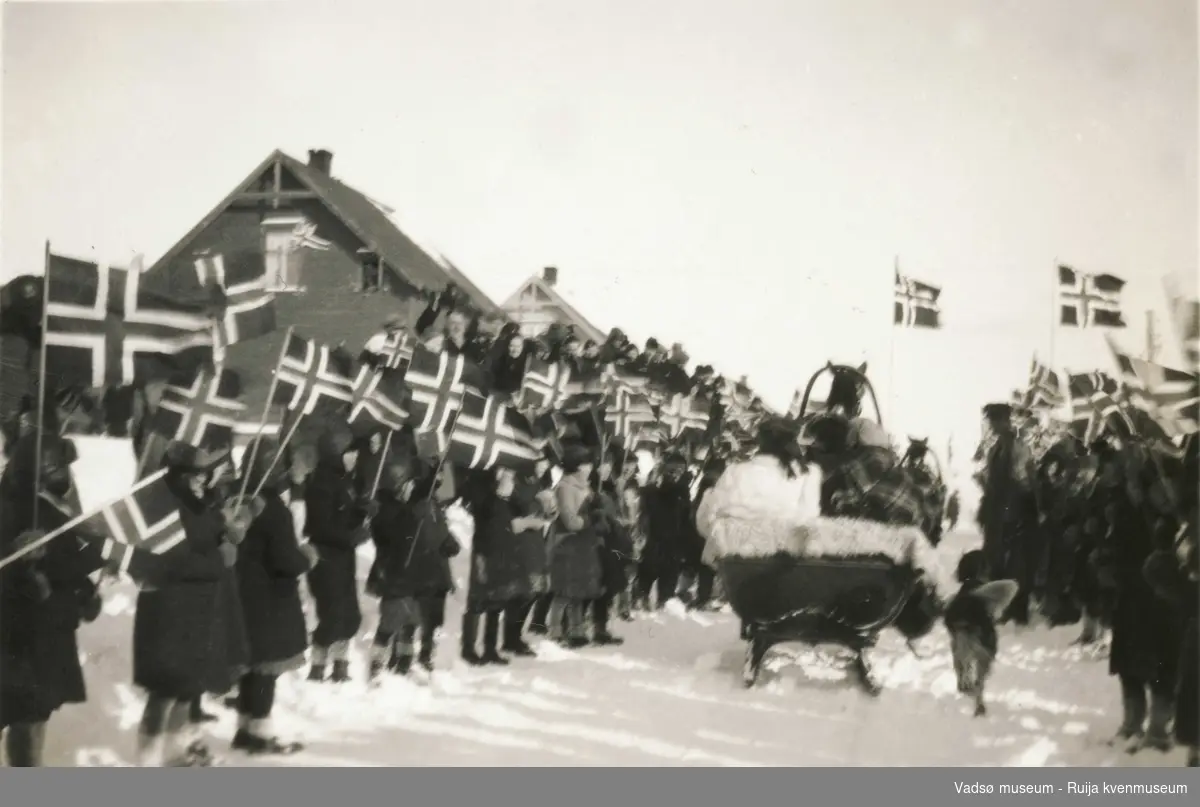 Sleden med de kongelige kjører gjennom folkemengde med flagg under kronprinsparets besøk i Vadsø i 1934.