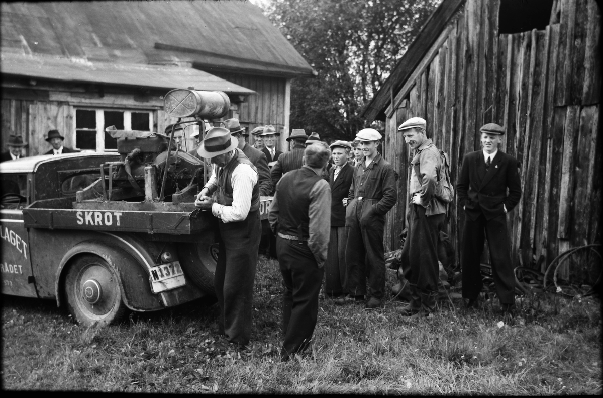 En skrothandlare besöker en gård och reglerar flakets sarg på bilen. Möjligen är det ett gengasaggregat han har på flaket. En samling män står och betraktar ekipaget.