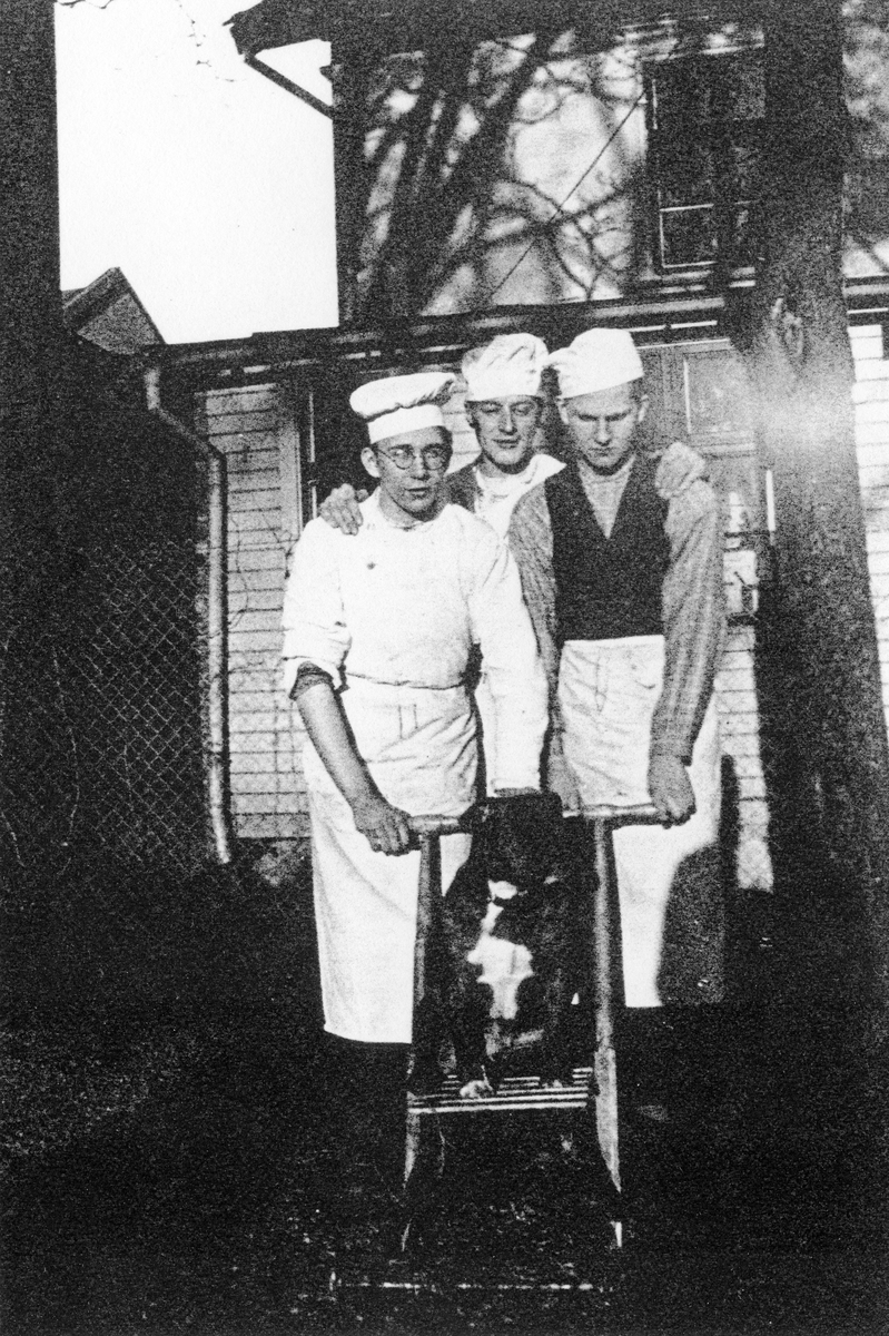 Tre manliga bagare på Ljungblads konditori står ute på gårdserveringen framför en spark med en hund som sitter ovanpå.