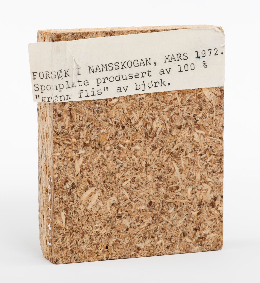 En rektangulær sponplate, vareprøve på 8,8 cm x 7 cm x 1,8 cm. Plata har glatt fram- og bakside.  På vareprøvas ene side er det påklistret en lapp med denne teksten: "Forsøk i Namskogan, mars 1972. Sponplate produsert av 100 % "grønn flis" av bjørk"