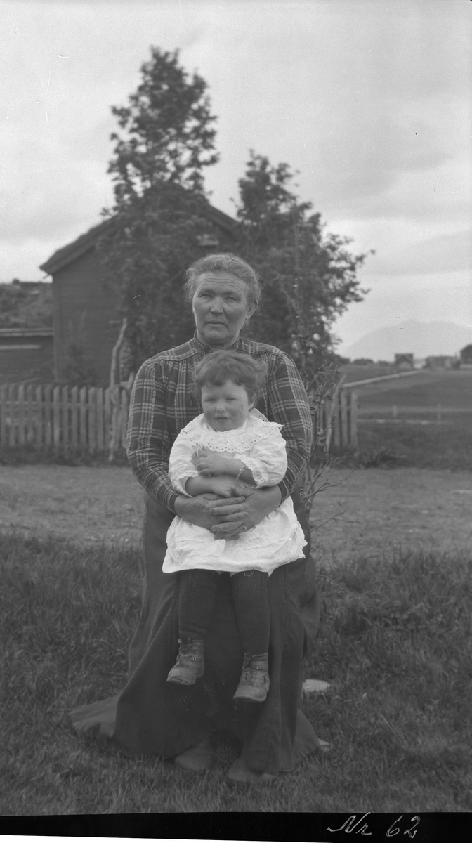 Portrett av eldre kvinne med barn på fanget sittende på gårdstun.