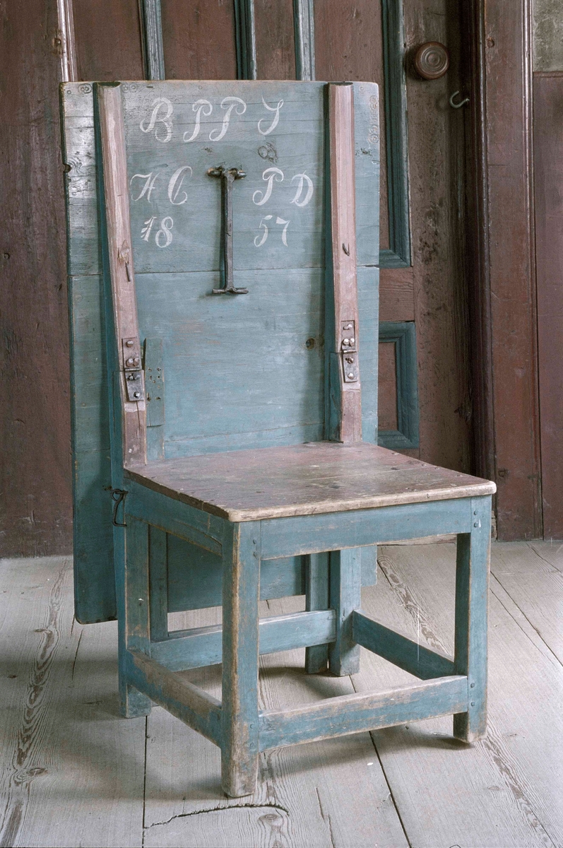 Bordstol av trä, rektangulär bordskiva bildar stolens rygg, ramslåar.
Då skivan är nedfälld stöder en järnfot mot stolens säte, utfällbar grind i stolens rygg. Sarg, ben och skivans underrede blåmålade, sits och ryggståndare röda.
Märkt: " B P P S / H C P D / 1857".
