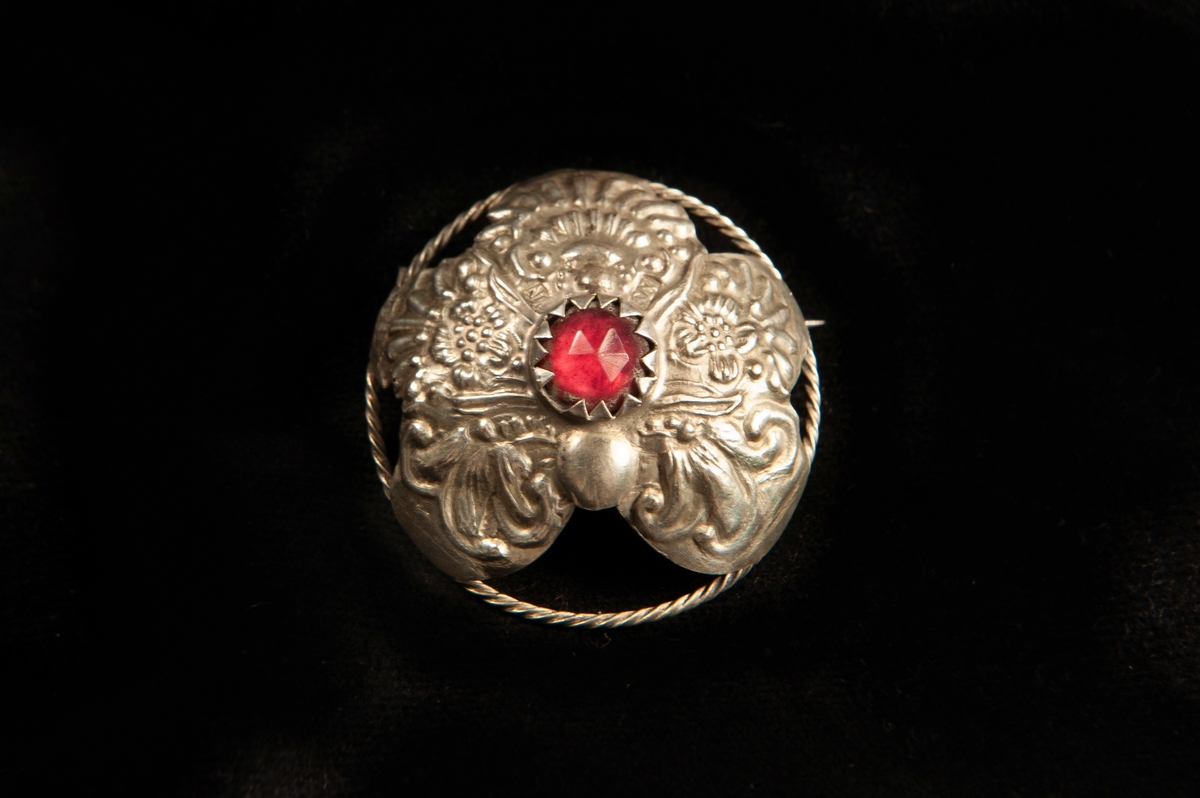 En rund malja av ciselerat och delvis genombrutet silver med fem "tungor" med blomdekor. En infattad sten av rött glas. Spännet, maljan, har gjorts om till en brosch.