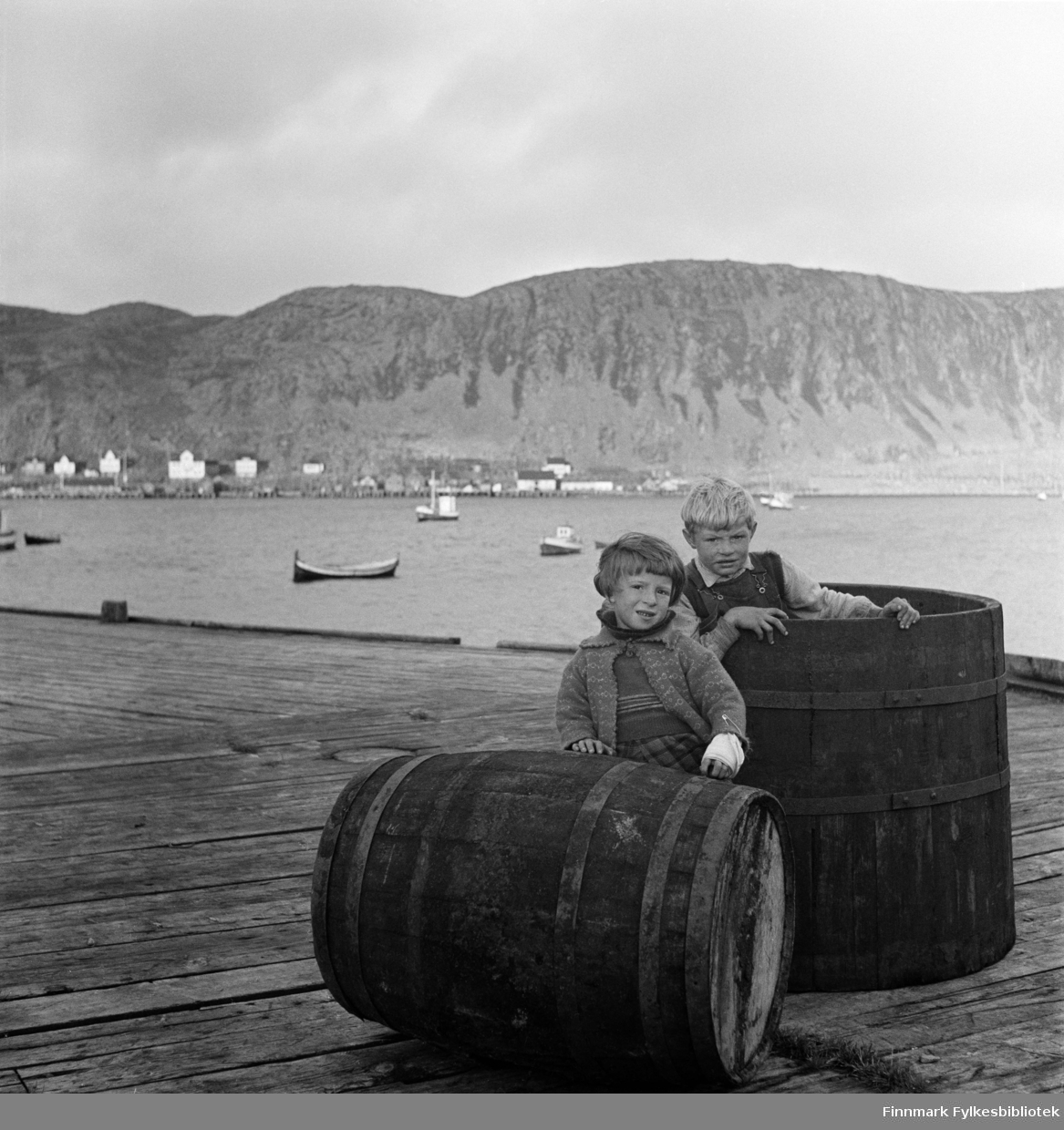 Åse Isaksen og Alf Nilsen (født 15.12.1933) på Nissen-kaia i Kjøllefjord, fotografert i 1940. Åse har bandasje på venstre arm med stor sikkerhetsnål. På andre siden av bukta sees flere fiskebruk og fiskemottak, hotell og boliger i Kjøllefjord. På fjorden ligger fiskebåter oppankret. En nordlandsbåt nærmest kamera. Åse har en strikket jakke og genser på seg og rutete skjørt eller bukser. Alf ser ut til å ha på seg snekkerbukser og skjorte. Sannsynligvis er dette på Nissenkaia.