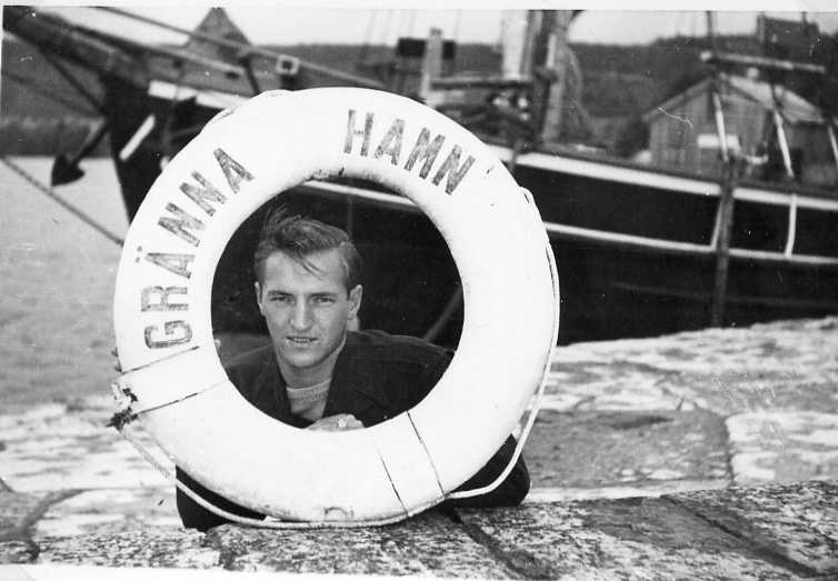 En ung man kikar ut genom en livboj märkt Gränna Hamn, sannolikt i Gränna hamn. En skuta ligger vid kaj i bakgrunden.