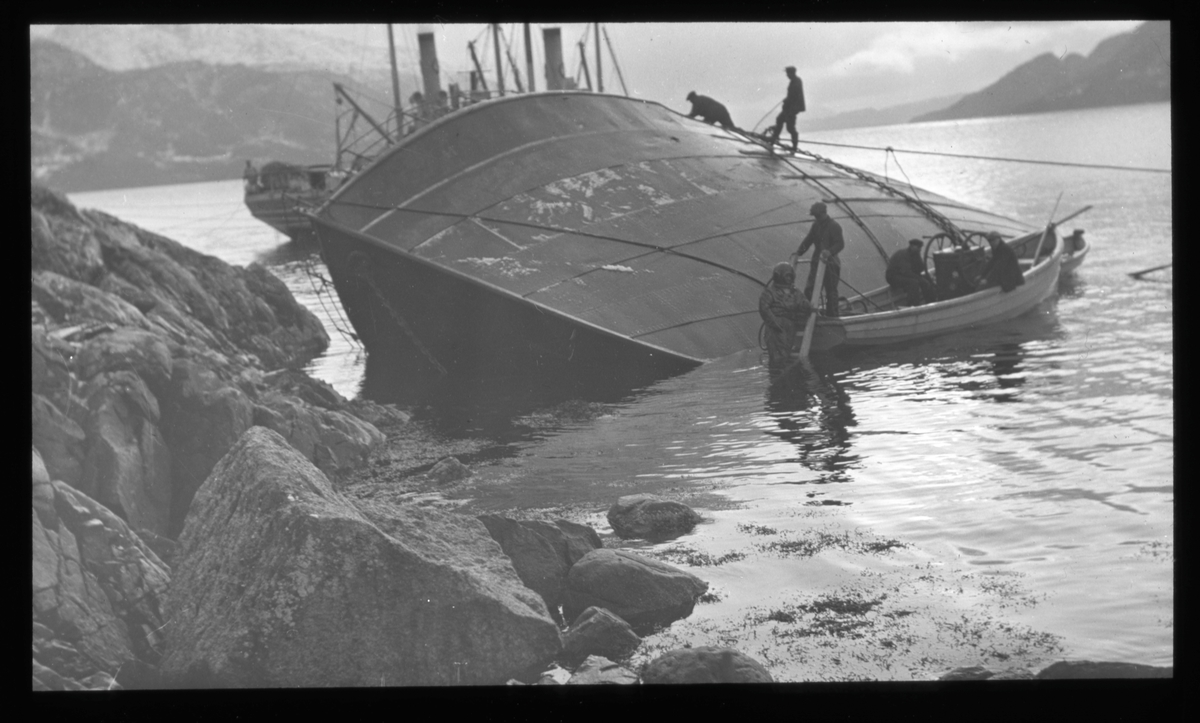 Berging av kantret skip fjæresteinene. Fire mann klatrer på skutesiden, liten lettbåt langs skutesiden.