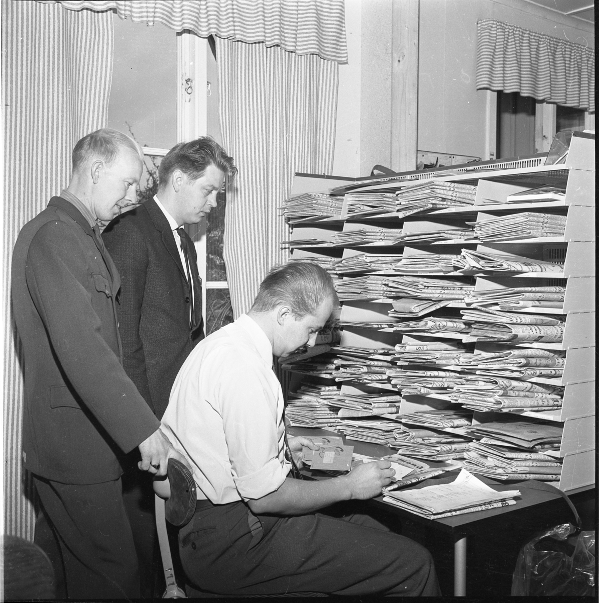 Premiär för lantbrevbärare i Gränna, 1 okt 1967. Henry Kvist och Rolf Lindén stående bakom Anders Hjelte som sitter i vit skjorta och slips vid ett bord med post i händerna. På bordet en hög hylla med postfack fyllda med tidningar.
