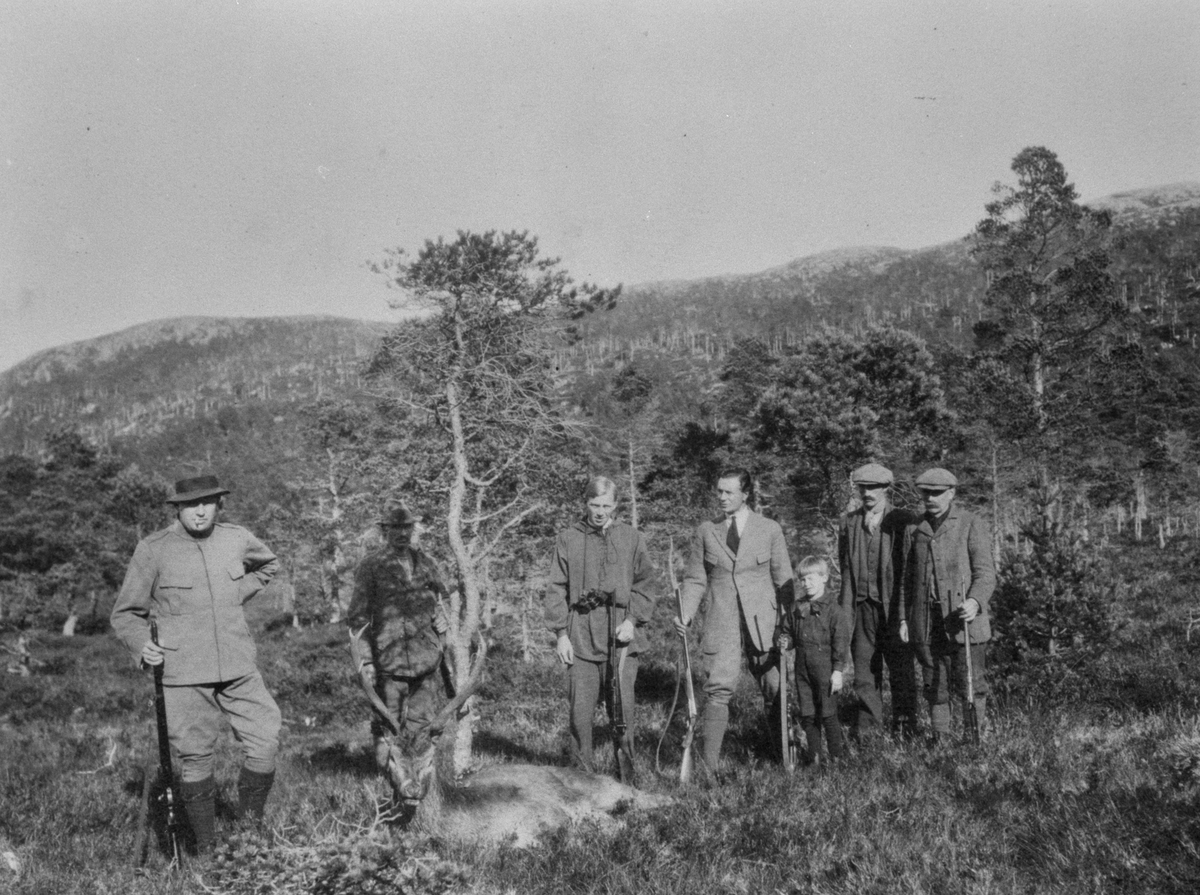 Gruppebilde med fremvisning av hjort utenfor Skogodden jakthytte, Strøm, Melandsjø. 7 menn og 1 gutt.
