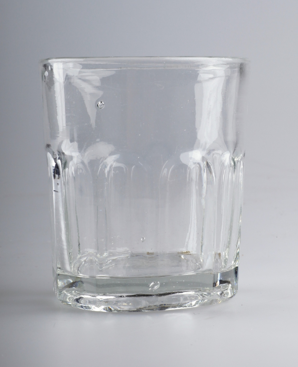 Munnblåst glass, blåst i form med flater som skal illudere slipning. Glasset er tykt og har mange ujevnheter og bobler i glassmassen. Bunnringen er slipt, slik at glasset står plant.