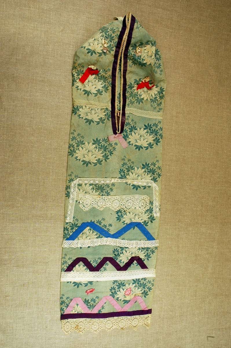 Dåpspose/hettepose i bomull med påtrykt bladmønster, dekorert med blonder, silkeband og stoffrosetter.