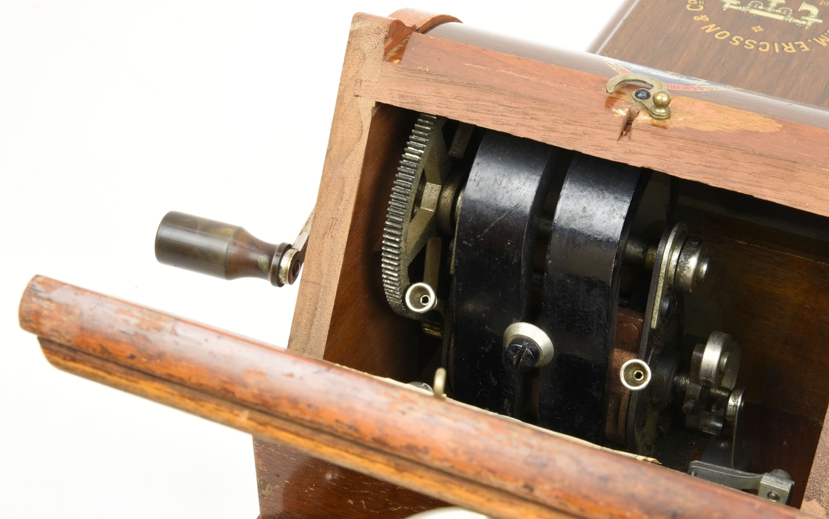 En väggtelefon av trä från tidigt 1900-tal gjord av L.M. Ericsson.

Mikrofonen är monterad direkt på telefonen och ser ut som en metalltratt, hörapparaten är handhållen och är kopplad till telefonen via en elkabel. På väggtelefonens överdel finns skruvbara mässingsfästen. Nedanför mikrofonen finns en ringklocka och ett svart påskruvat metallhölje med påmålad ornamentik. Mitt på väggtelefonen sitter en utstickande trälåda som innehåller mekaniska komponenter som rör på sig när man rör på den vev som sticker ut ur lådans högra sida. På undersidan av locket till trälådan finns en pappersetikett med en förteckning över telefonen. Nedanför trälådan finns ett hyllplan som täcks av en metallkåpa (med målad träimitation), i det utrymmet ska det finns ett batteri.