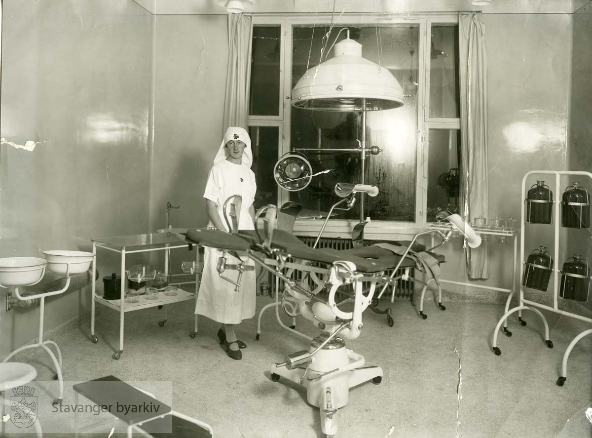Sykepleier på gynekologisk avdeling. Gynekologstol i midten av bildet. Annet utstyr og apparater rundt om i rommet. .Avlevert av Harald Bjørnestad.