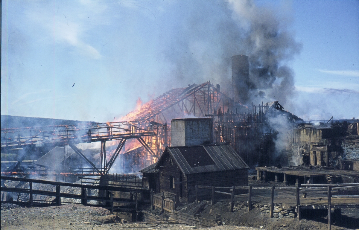 Brann i smeltehytta på Røros, lørdag 19. juli 1975, sett fra østsida av elva. Foran i bildet sees øvre maskinhuset og sleggbanen. Lenger bak brenner det i kølbrua