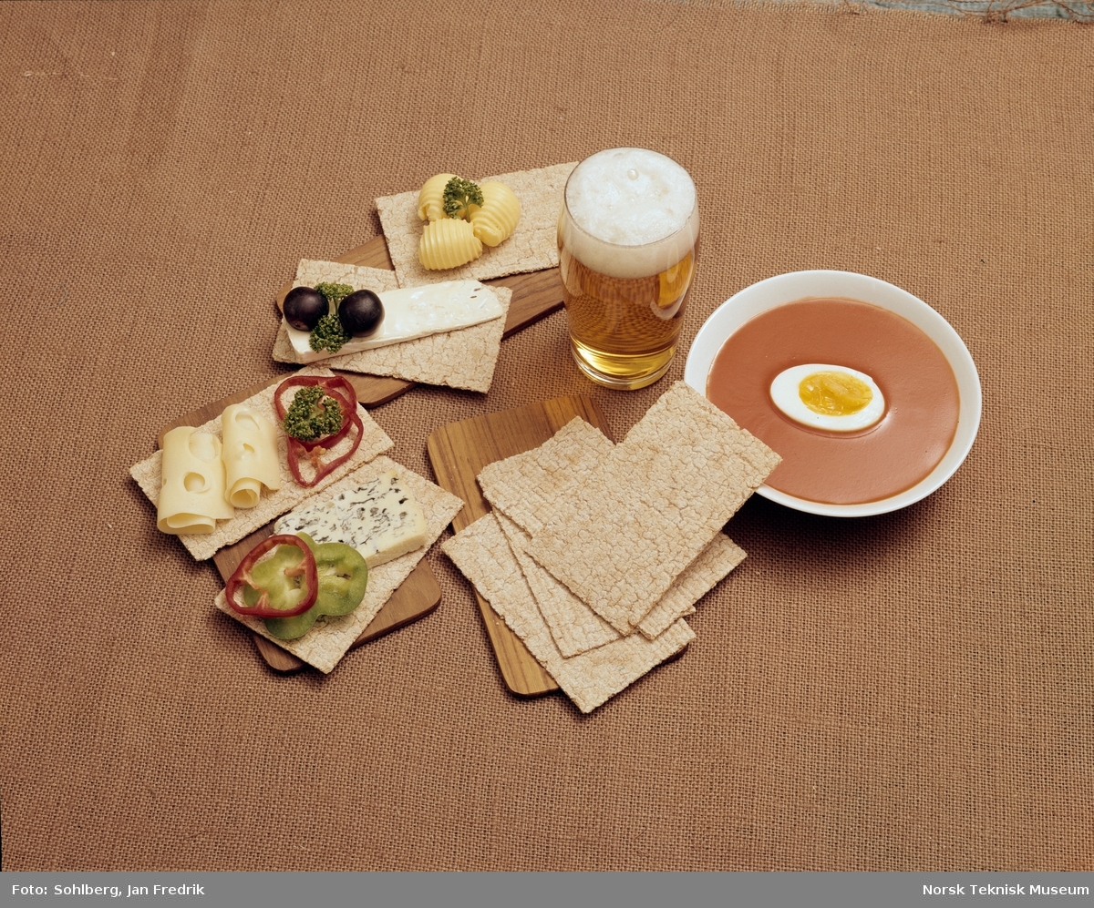 Bildet reklamerer for flatbrød av merket Korni. Et bord er dekket med tomatsuppe og spekemat med flatbrød til. Et glass med skummende øl står sammen med maten.