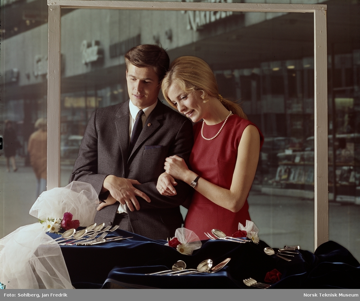 Et moteriktig kledd ungt par som ser på sølvtøy. De har ringer på fingrene. Kanskje et nyforlovet par som ser på bryllupsgaver?