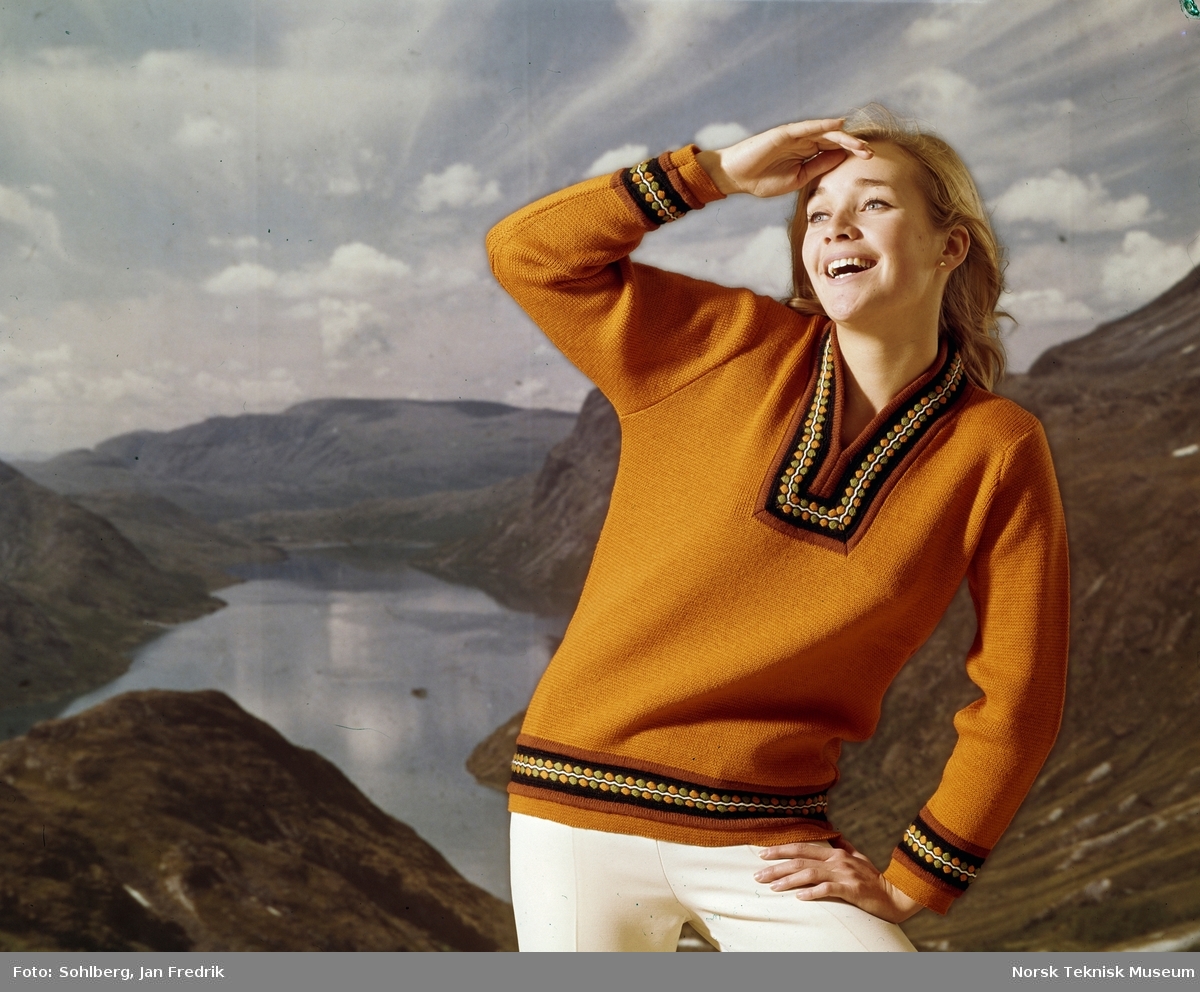 En kvinnelig modell viser fram en okerfarget strikkegenser. Studiofoto med bakgrunn av norsk natur, fjellheim og fjord.