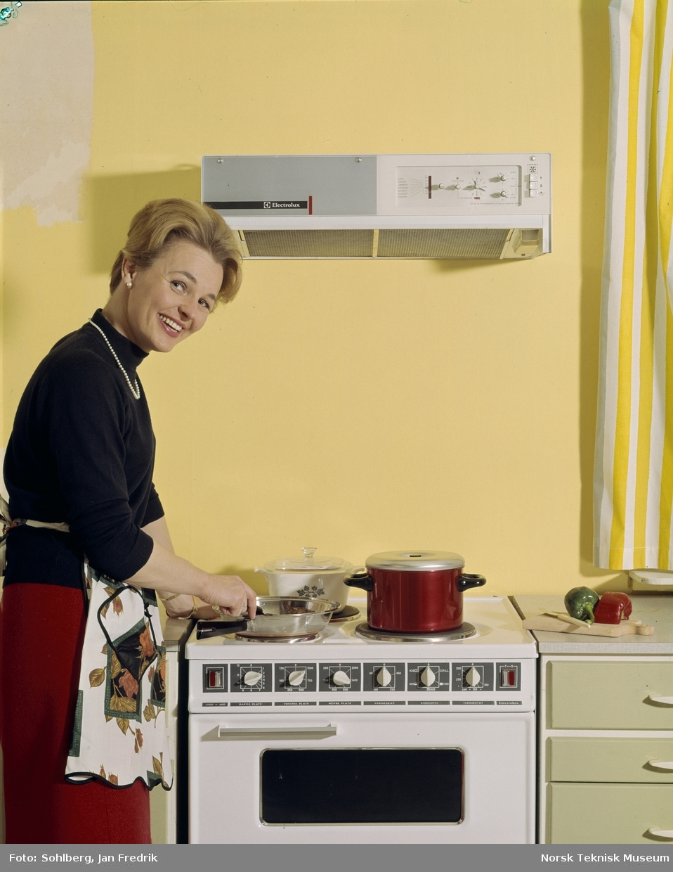 Reklamefoto for komfyr og vifte fra Elektrolux. Med moderne kjøkkenutstyr er livet som husmor ganske enkelt. En smilende kvinnelig modell lager mat ved komfyren.