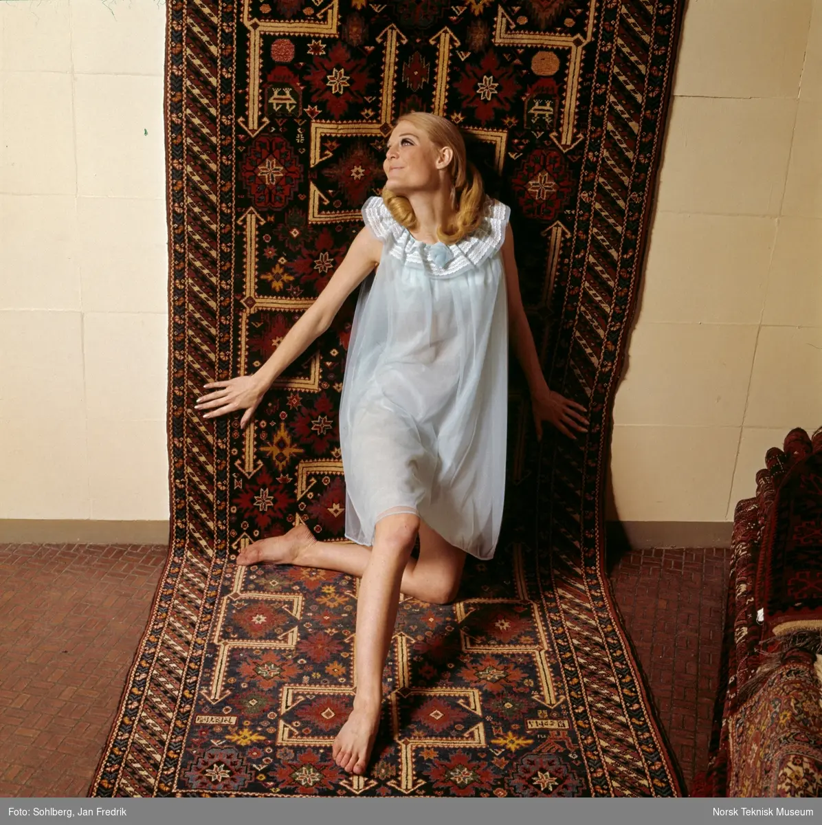Tidlig norsk motefotografi. Kvinnelig modell poserer i nattkjole foran et orientalsk teppe.