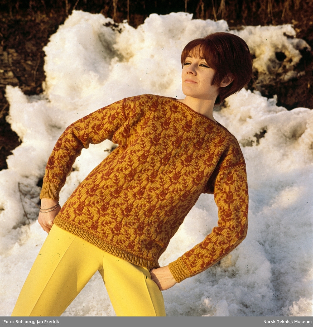 Strikkemote fra 1960-tallet. Kvinnelig modell viser en oransje og gul strikkegenser med mønster. Fotografert utendørs i vinterlandskap.