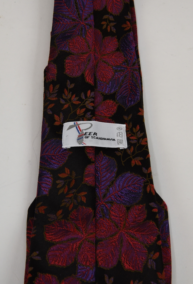 Mönstrad slips med svart botten. Mönstret består av broderade blommor och lövverk i bland annat violett och rosa, mönstret upprepas längs hela slipsen. Slipsen är fodrad med en röd textil med grå text.