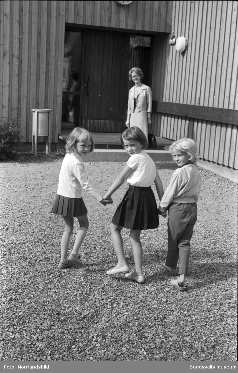 Skolstart i Skönsbergs skola (Hellbergsskolan). Tre små flickor möter sin fröken vid en av de tre, sedermera rivna,  lågstadiepaviljongerna utmed Bruksgatan.