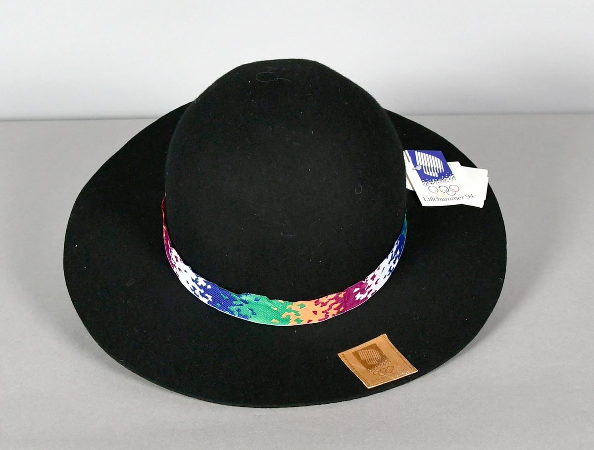 Svart hatt av vadmel med brem og bånd med krystallmønster i hvitt, blått, grønt, rosa og lilla. Skinnmerke på bremmen med logo for Lillehammer '94.