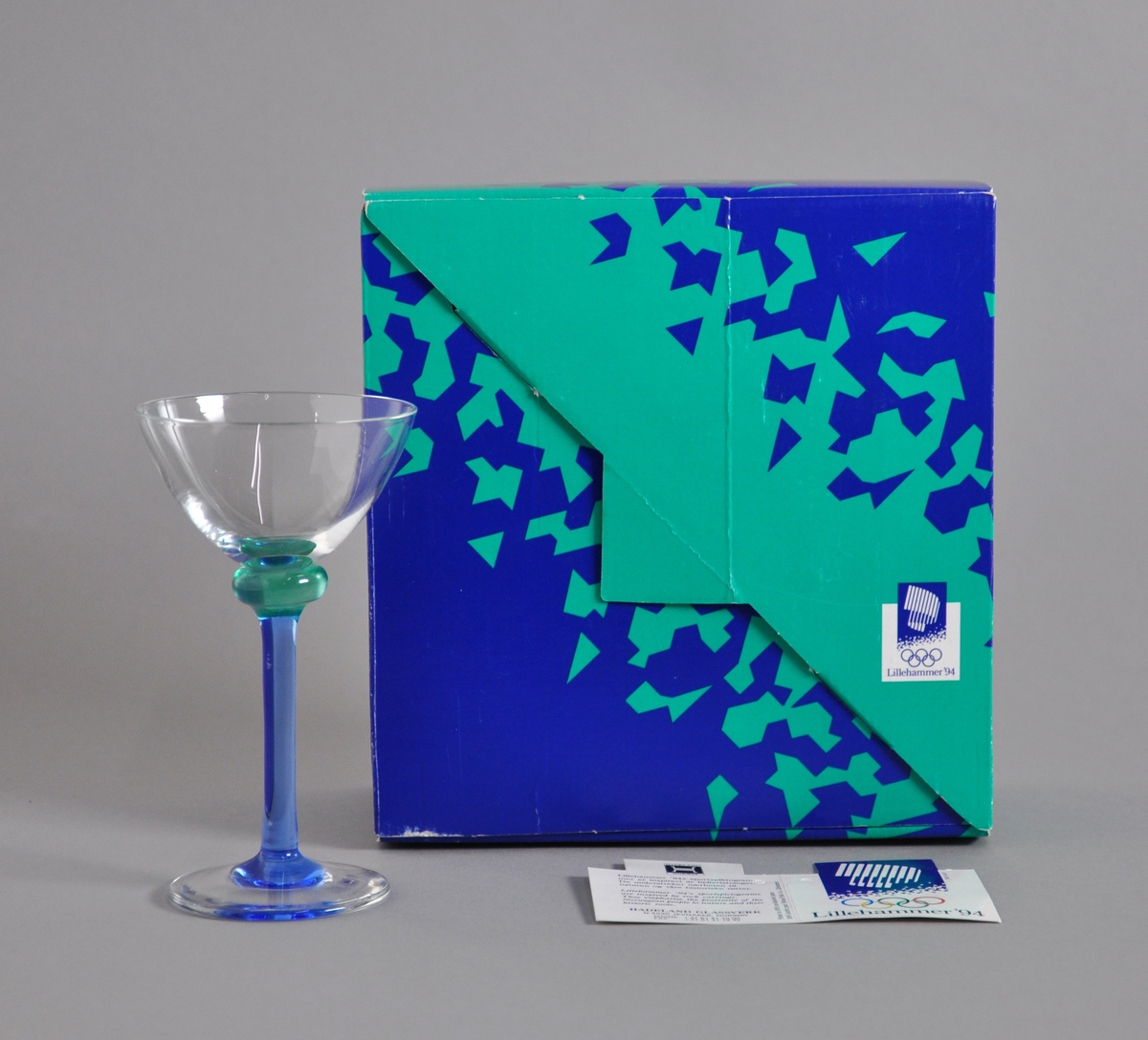 Likørglass med blått stett med fakkelmann, turkis kant like under glasset. Glasset ligger i original emballasje.