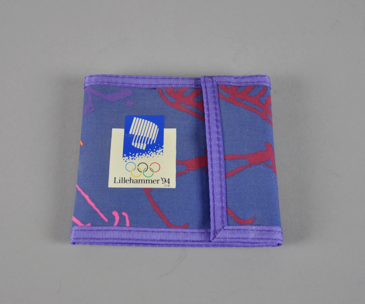 Blå lommebok med lilla kantbånd. Piktogrammer i flere farger. En lomme med glidelås, tre lommer for kort og lomme for sedler. Lommeboken har borrelås og emblem for Lillehammer '94 på forsiden.