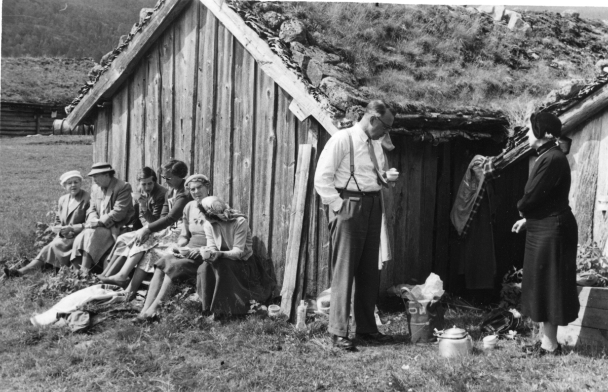 Presten i Suldal og en gruppe kvinner drikker kaffe bak naustene i Kvilldal. De er på utflukt fra Lindum. På bilde 2 står Frida Johannessen og snakker med presten. På bilde 3 sitter Frida Johannessen til venstre, mens presten holder kaffekanna til høyre.