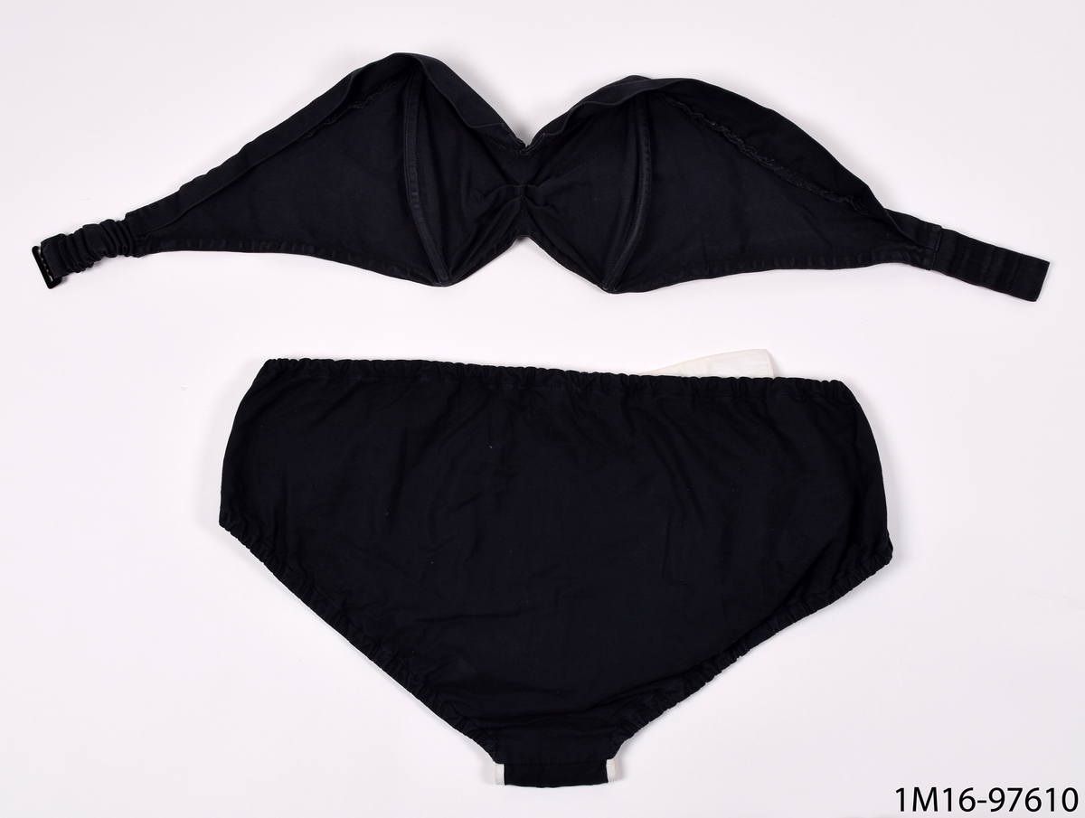 Baddräkt, bikini av svart maskinvävd bomull, med stålfjädrar. Bikini byxorna har ett vitt skärp i midjan samt en vit kant vid grenen framtill.