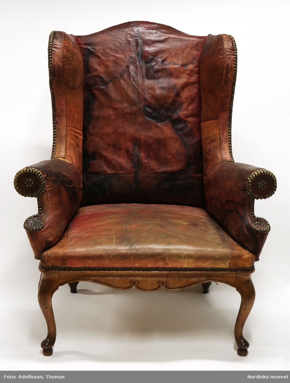 Fåtölj med "öronlappar", även kallad sovstol, rokoko, omkring 1750, sannolikt tillverkad i Stockholm efter engelsk modell. Helt klädd med rött skinn (marokäng/saffian), fläckat och kraftigt blekt. Skinnklädseln fästad med tätt sittande möbelspik av mässing. Underrede av brunbonad (ursprungligen rödfärgad) al. Ryggens insida har stora, mörka fläckar. 
Anm: Renoverad. Sitsen moderniserad med resårstoppning. Bakbenen utbytta mot ben av ek. Sliten skinnklädsel.
/Anna Arfvidsson Womack 2021