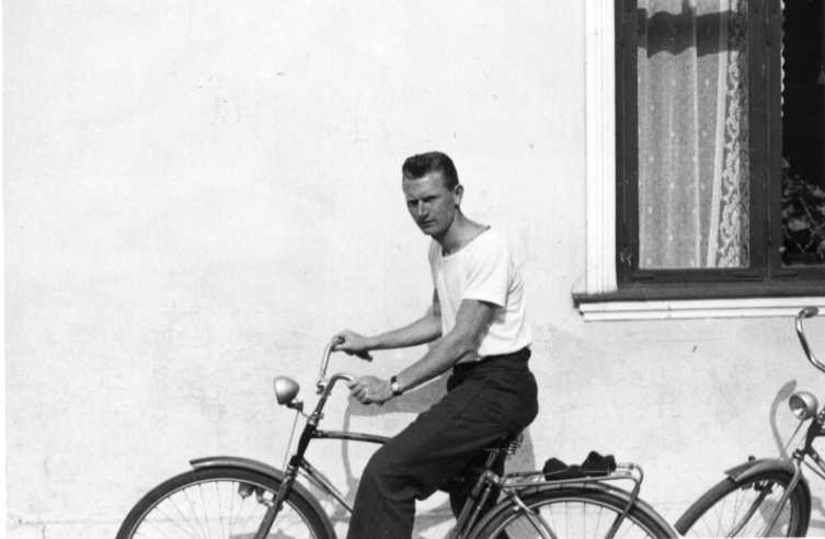 En man i t-shirt sitter på en cykel vid en ljusputsad husvägg.