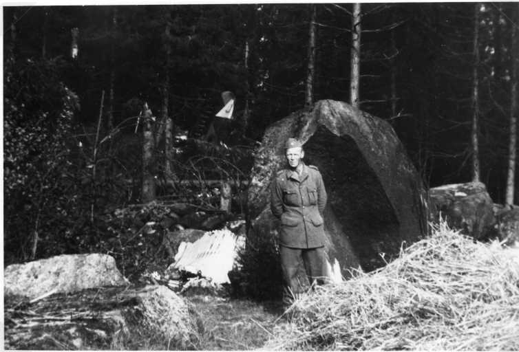 En svensk militär i uniform framför en stor sten och bakgrunden stjärtpartiet av ett kraschat bombplan.