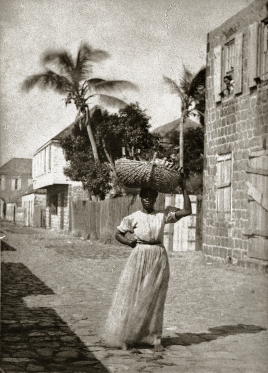 Gatubild från Gustavia. 
Kvinna med korg på huvudet. Ett barn tittar ut från ett fönster.