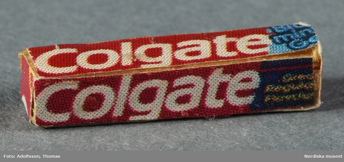 Två tandkrämstuber av märket Colgate. Den ena a) är av vitmålad avrundad metall med röd etikett och den andra b) föreställer en pappkartong och är förmodligen tillverkad av trä och papper.