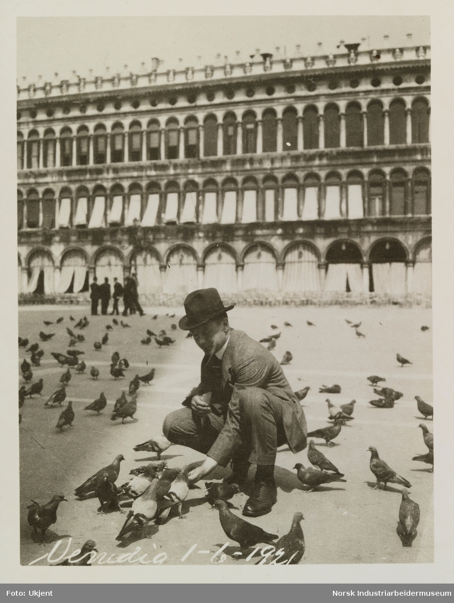 Mann sitter på huk med duer på åpen plass i Venezia