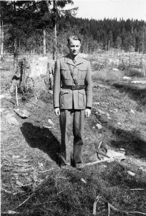 En ung man i militär uniform stående i skog framför en gärsgård.