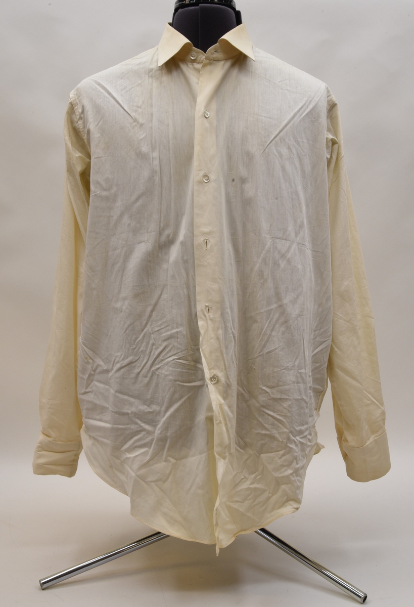 Långärmad skjorta av vit bomull. Skjortan har en enradig knäppning med sju knappar av plast av imiterad pärlemor. Kragen har en kragplatta av genomskinlig plast inuti kragsnibbarna och är märkt på insidan med både stämplar och lapp.