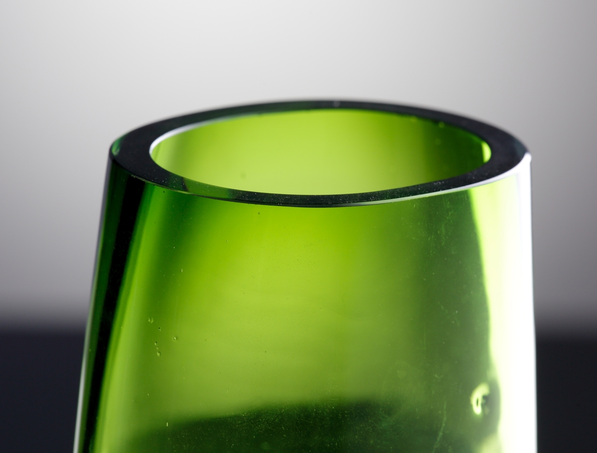 Oval och konisk till formen.
Grönt underfång med tjock ofärgad botten.
På bottens sidor två intryckta markeringar.
Vasen är av sekundakvalité och ofärdig i det att den kullriga bottnen inte planslipats.