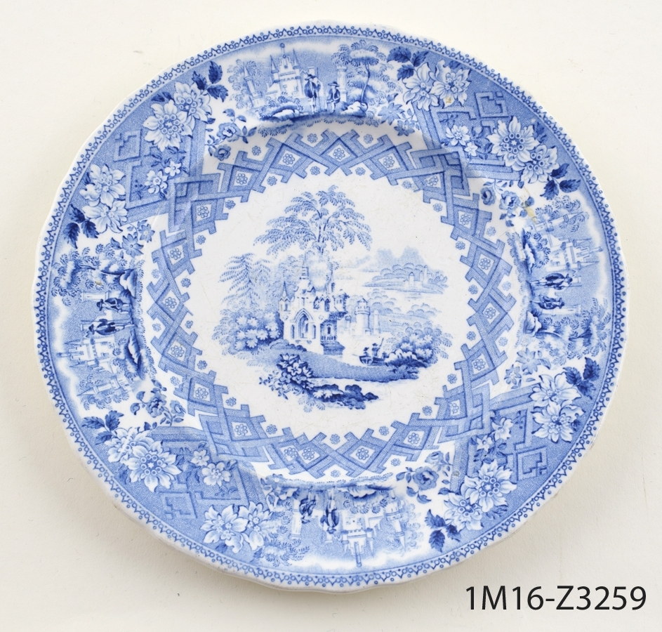 Rund vit assiett med tryckt blå dekor föreställande (mittmotiv) landskap och ornamenterad bård på bräm och kepkant.