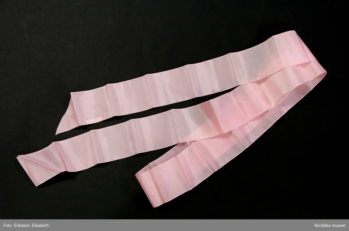 Dopklänning (a) av tunt vitt syntettyg med stråveck, två rosa rosetter av blankt kontsidentyg upptill, mitt fram på livet, liksom en maskintillverkad spets, påsydd i v-form. Spetskant med udd nedtill på klänningen. Korta puffärmärmar, ihopdragna med rosetter av rosa konsidentyg. Knäppning upptill mitt bak, omlottsprund med tre små vita plastknappar.
Till dopklänningen hör ett band (b) av rosa konstsidentyg att knyta kring livet.
/Helena Lindroth 2019-02-20