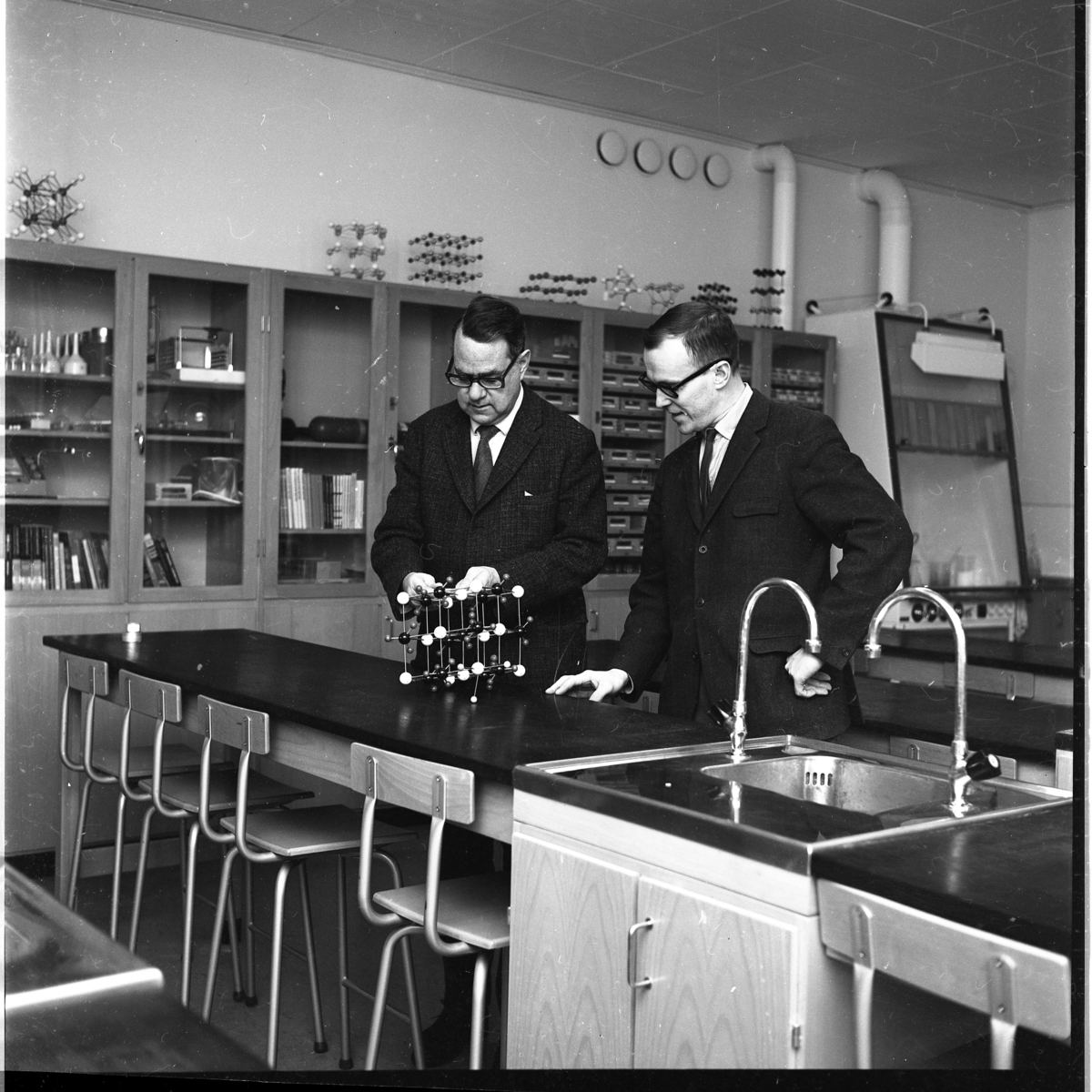 Två män står vid en lång skolbänk som separeras av en diskho. De står framför en modell av en molekyl. Utmed väggen i bakgrunden står höga skåp med glasdörrar. Det är rektorn Göte Östlund och ämnesläraren Hans Fridell till höger som befinner sig i en lärosal på Ribbaskolan.
