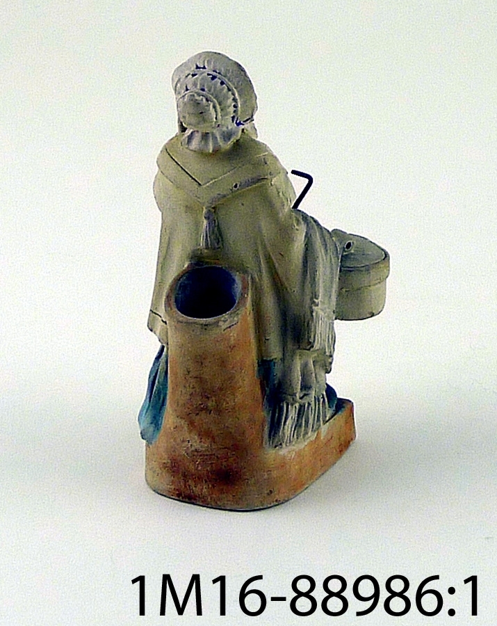 Figurin föreställande kvinna som bär på korg, väska, paraply och liten hund. Figurinens utförande har drag av karikatyr.