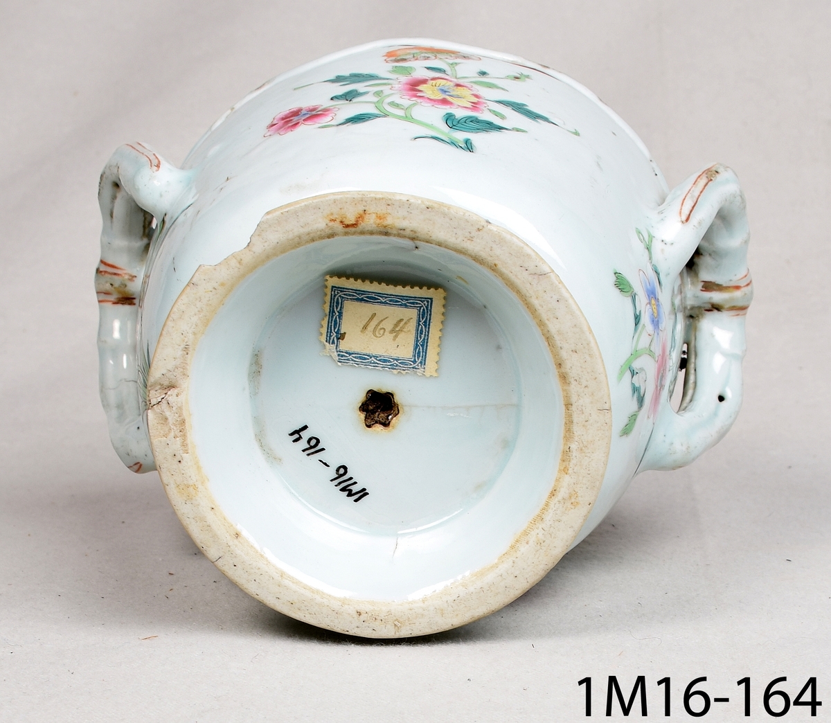 Rund kruka av keramik med ljusblå glasyr och handmålad blomdekor i många färger och guld. Krukan har två hänklar i överkant och en vågformad kant.