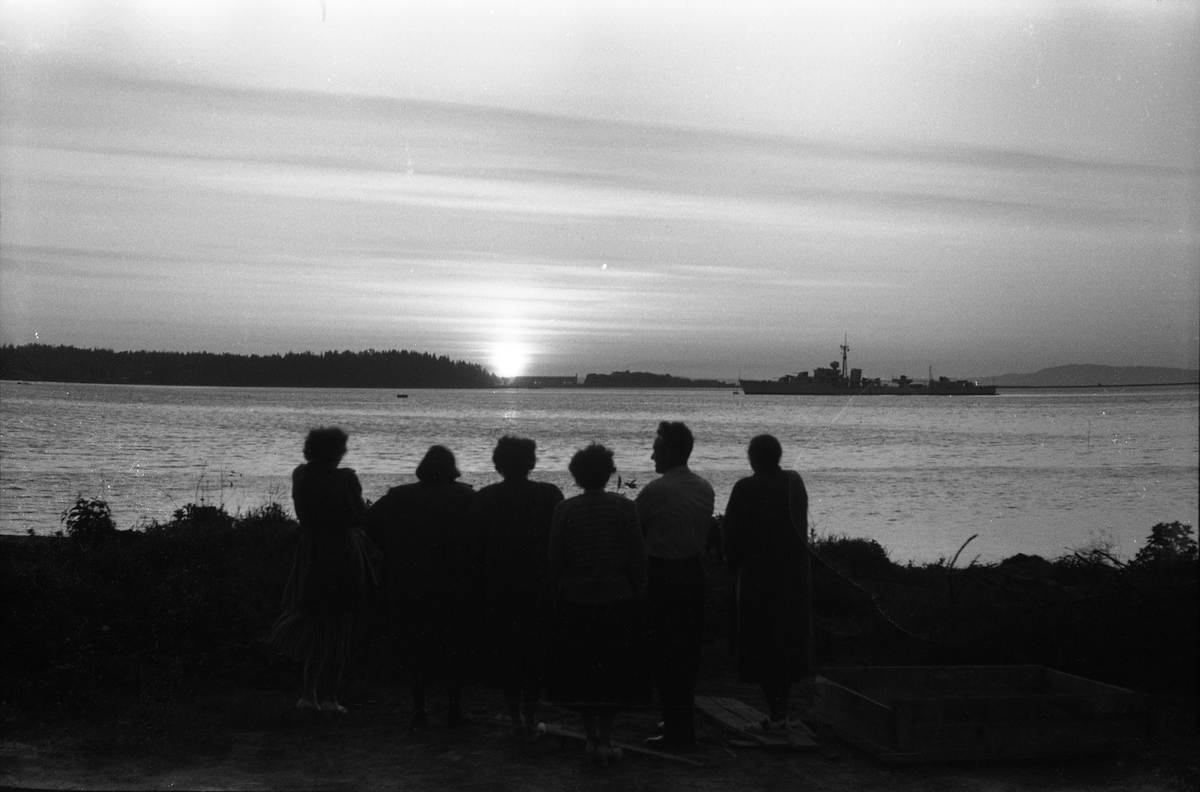 To avfotograferte bilder av ei gruppe personer samlet ved en fjord i solnedgang eller muligens midnattsol. Et fartøy passerer ute på fjorden.