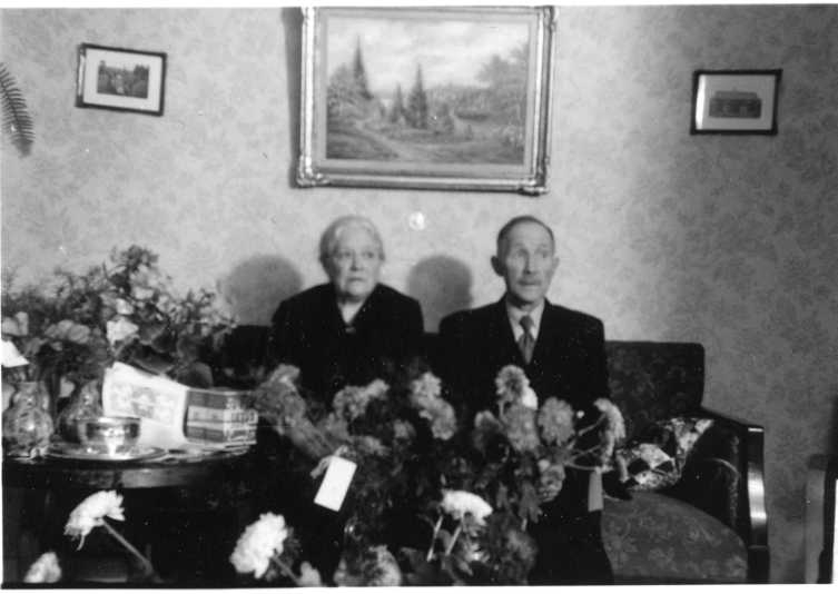 Anna och Teodor Johansson sitter i en soffa. Till vänster ett runt bord med blommor och även blommor framför dem. På väggen bakom dem hänger en större tavla med en mindre på vardera sida. Möjligen firas en födelsedag eller annan högtid.